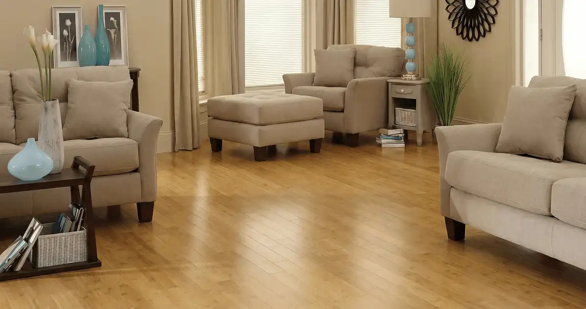 A living room with professionally installed bamboo flooring, beige sofas, a matching ottoman, a side table with a blue lamp, vases, framed pictures, and large windows with blinds and curtains in Raleigh, NC